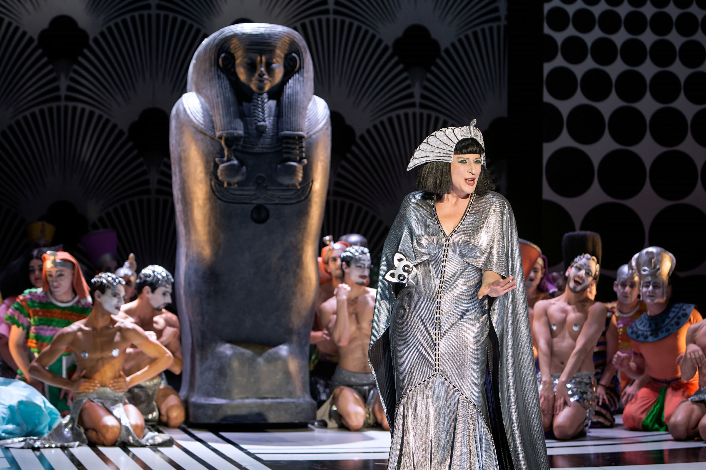 Dagmar Manzel als Cleopatra auf Bühne Komische Oper Berlin, im Hintergrund ein Sarkophag, umgeben von Tänzer:innen sitzend