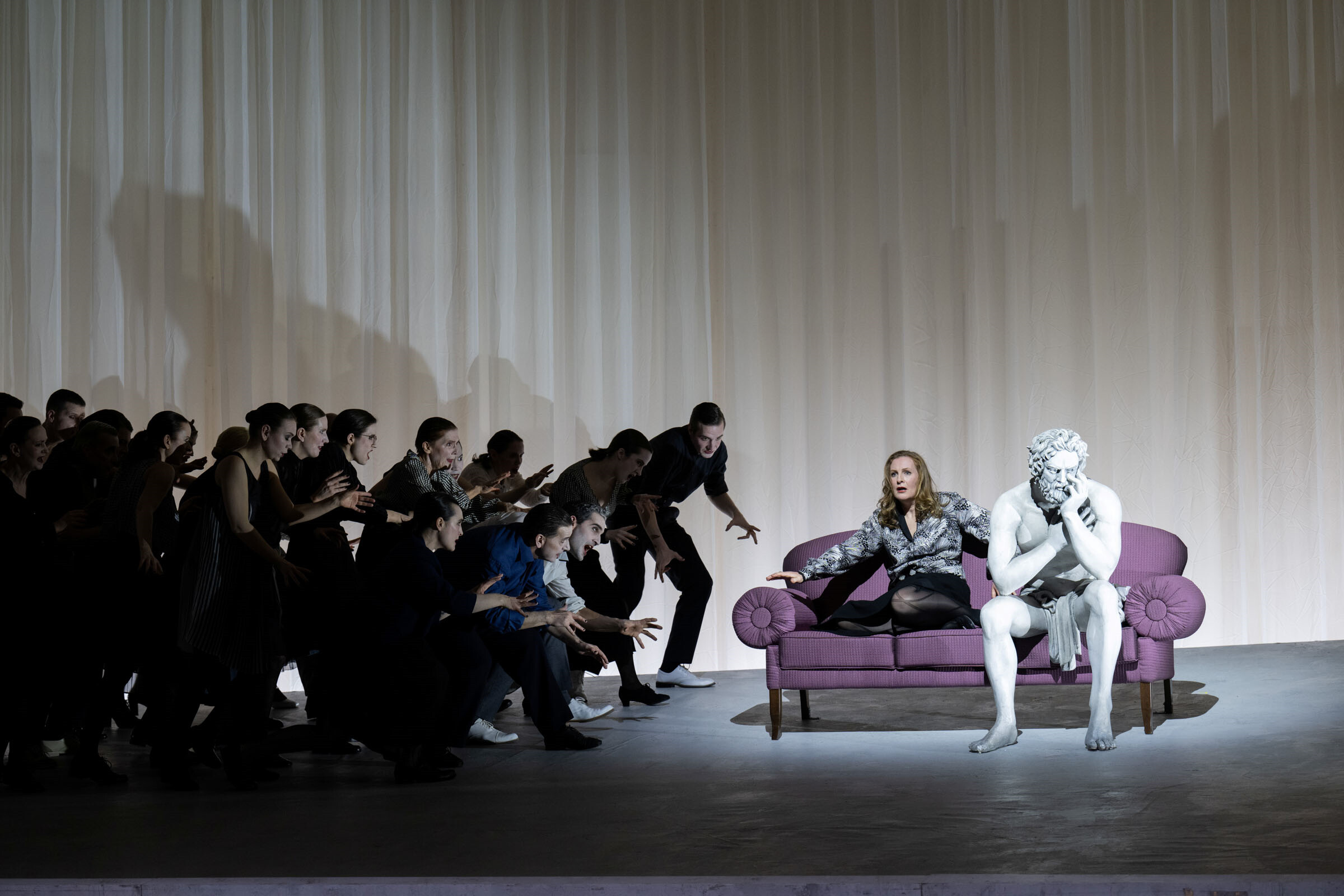 Paula Murrihy als Dejanira neben Statue des Hecules auf rosa Sofa sitzend, links Gruppe von TänzerInnen aus sie zu laufend
